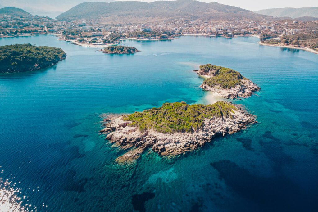 Vista aérea de Ksamil: el Caribe albanés, una zona costera con varias islas pequeñas y verdes rodeadas de aguas cristalinas y un pueblo al fondo.