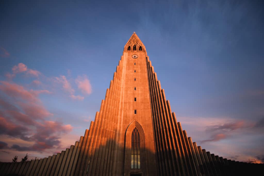 La iglesia más famosa de Reykjavik. Una fachada de iglesia de hormigón alta y puntiaguda con un reloj cerca de la parte superior, iluminada por la luz del atardecer contra un cielo con nubes dispersas. 
