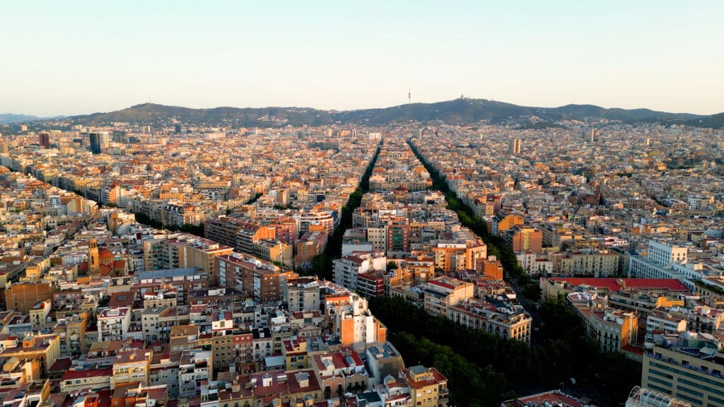 Vista aérea de un paisaje urbano en expansión con calles en forma de cuadrícula y numerosos edificios, muy similar a un MAPA turístico de Cataluña, con colinas distantes bajo un cielo despejado.