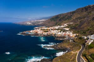 Ciudad costera con edificios blancos a lo largo de una costa rocosa, rodeada de colinas bajo un cielo azul claro. Se ve una carretera sinuosa en primer plano.