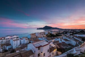 Un pueblo costero con edificios blancos al atardecer, con vistas al mar y una montaña lejana bajo un cielo colorido.