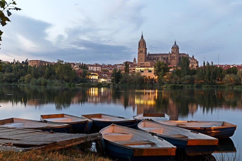 Pequeñas embarcaciones están amarradas cerca de un muelle de madera en un río. Al fondo, una catedral histórica y los edificios circundantes se iluminan al anochecer y se reflejan en las tranquilas aguas.