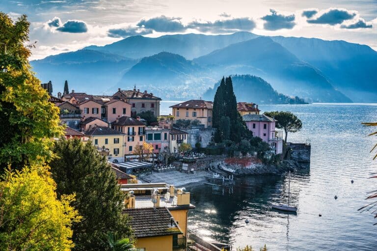 Vista panorámica de un pueblo junto al lago con casas coloridas, colinas y montañas distantes bajo un cielo nublado.