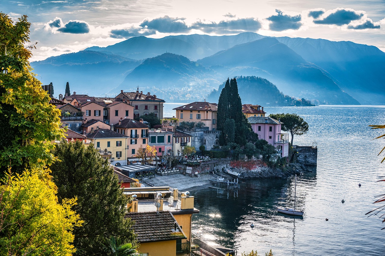 Vista panorámica de un pueblo junto al lago con casas coloridas, colinas y montañas distantes bajo un cielo nublado.