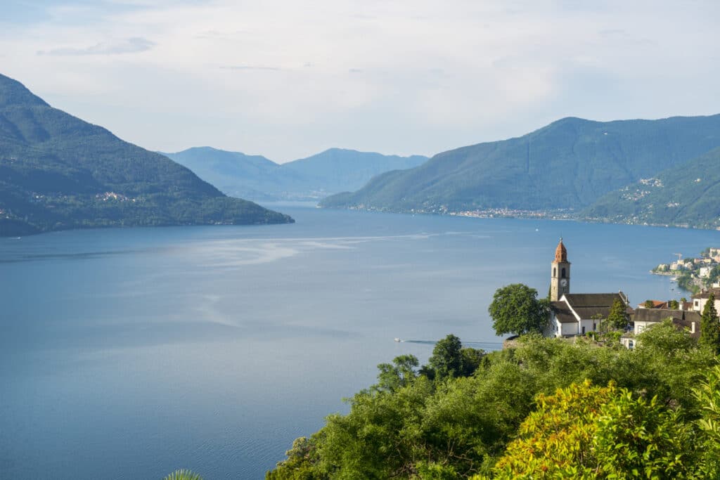 Una vista panorámica de un lago rodeado de montañas, con una iglesia y un pueblo en primer plano bajo un cielo parcialmente nublado.