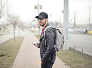 Hombre con una chaqueta de cuero y gorra de pie en una acera, sosteniendo un teléfono inteligente y llevando una mochila.