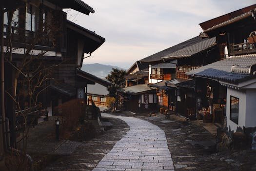 Estrecho camino de piedra que serpentea a través de un pueblo japonés tradicional con edificios de madera a ambos lados bajo un cielo nublado.