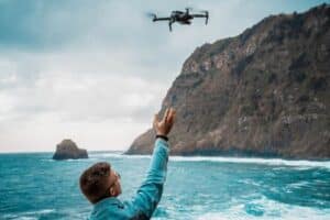 Una persona controla un dron sobre el océano cerca de un acantilado rocoso bajo un cielo nublado.