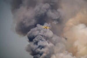 Un avión amarillo vuela sobre un humo espeso y oscuro que se eleva hacia el cielo, posiblemente procedente de un incendio que se encuentra más abajo.