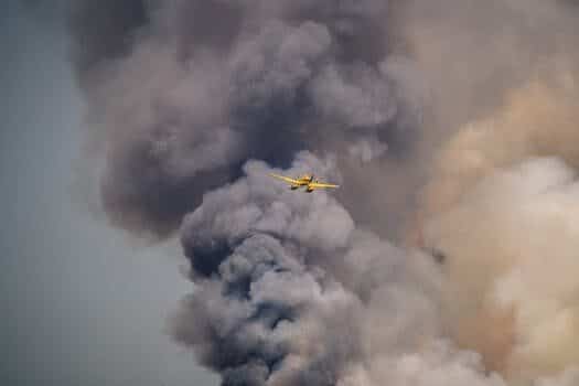 Un avión amarillo vuela sobre un humo espeso y oscuro que se eleva hacia el cielo, posiblemente procedente de un incendio que se encuentra más abajo.